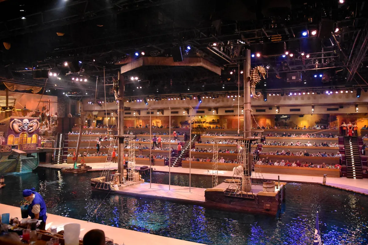 A vibrant setting showcasing a live water-based performance in a pirate-themed arena. Performers, a pirate ship stage, and an engaged audience create a sense of spectacle and immersive adventure.