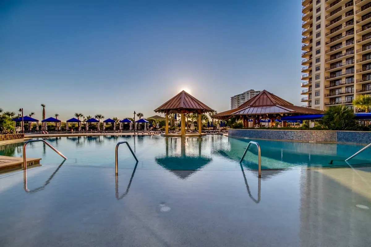 A luxurious outdoor pool area featuring clear blue water, comfortable cabana structures, and sunlight reflecting off the pool's surface. The tranquil setting is perfect for group relaxation and post-golf unwinding.
