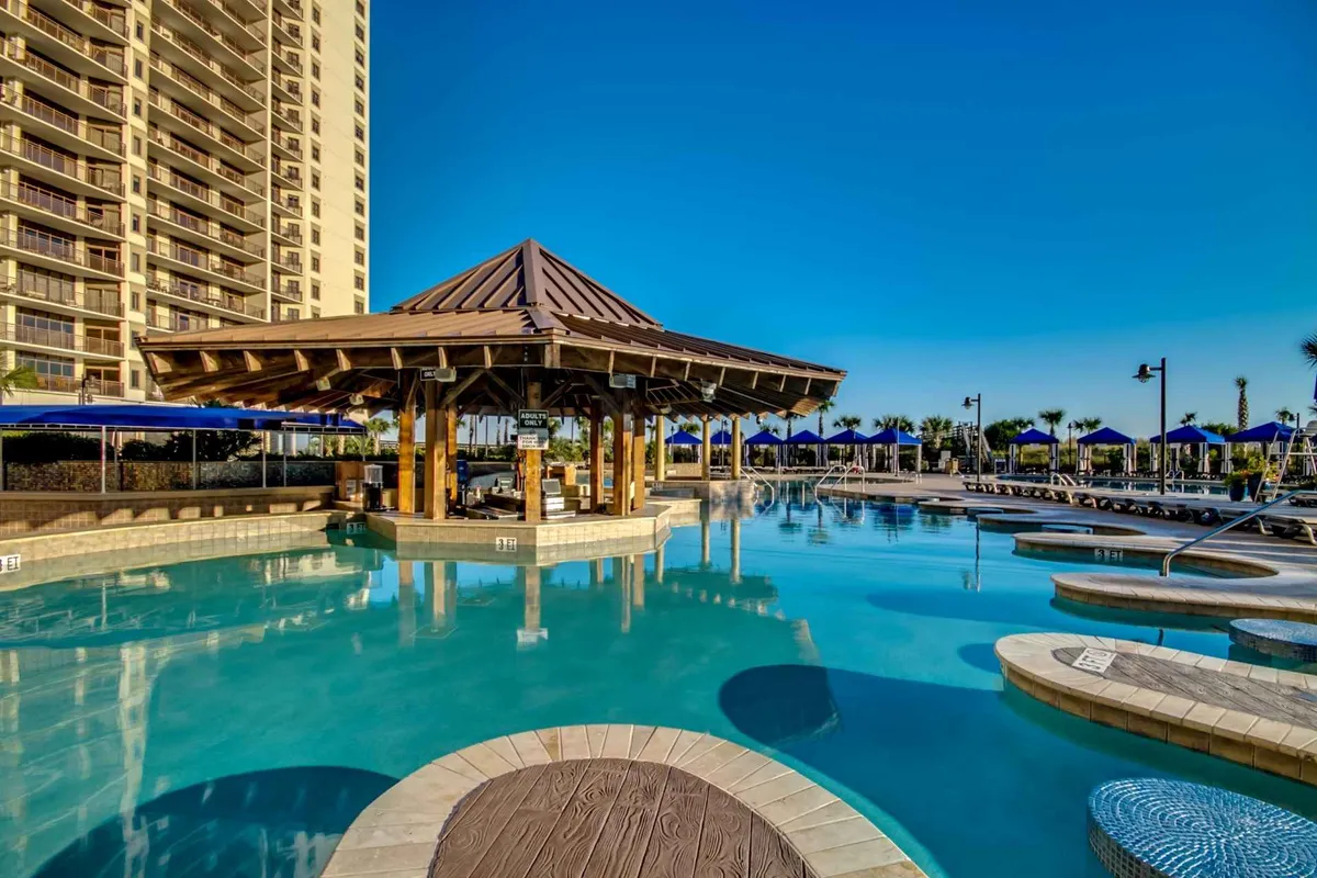 A vibrant exterior scene featuring a large resort-style pool with a cabana, plenty of lounge chairs, and a modern building in the background. Guests can be seen enjoying the sun by the pool, swimming, or relaxing with drinks under shaded cabanas.