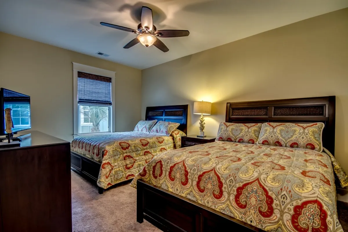 A relaxing bedroom with two beds featuring decorative bedding, a ceiling fan, and a lamp on the nightstand. A window with blinds lets in natural light. The space is ideal for families or friends traveling together, offering both comfort and flexibility.