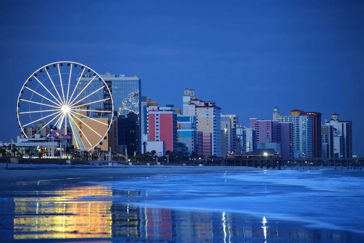 A lively view of a Ferris wheel lit up against the evening sky with colorful buildings and shimmering ocean reflections. The setting is playful and energetic, evoking the spirit of beachside amusement and nightlife.