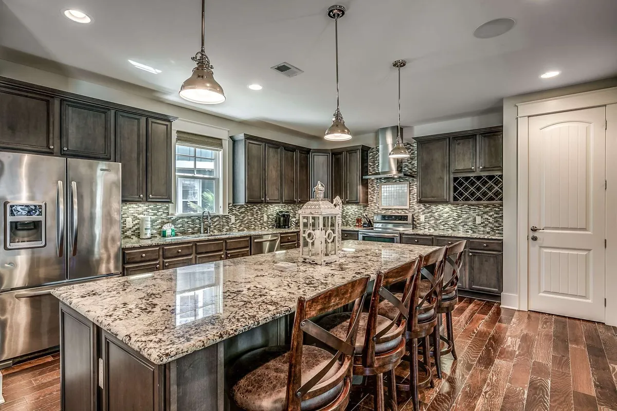 A warmly lit kitchen featuring a large granite island with stylish pendant lighting overhead. Stainless steel appliances, including a refrigerator and oven, are seamlessly integrated into dark cabinetry. Decorative touches like a lantern and neatly arranged kitchenware suggest a welcoming and functional space perfect for meal prep and entertaining.