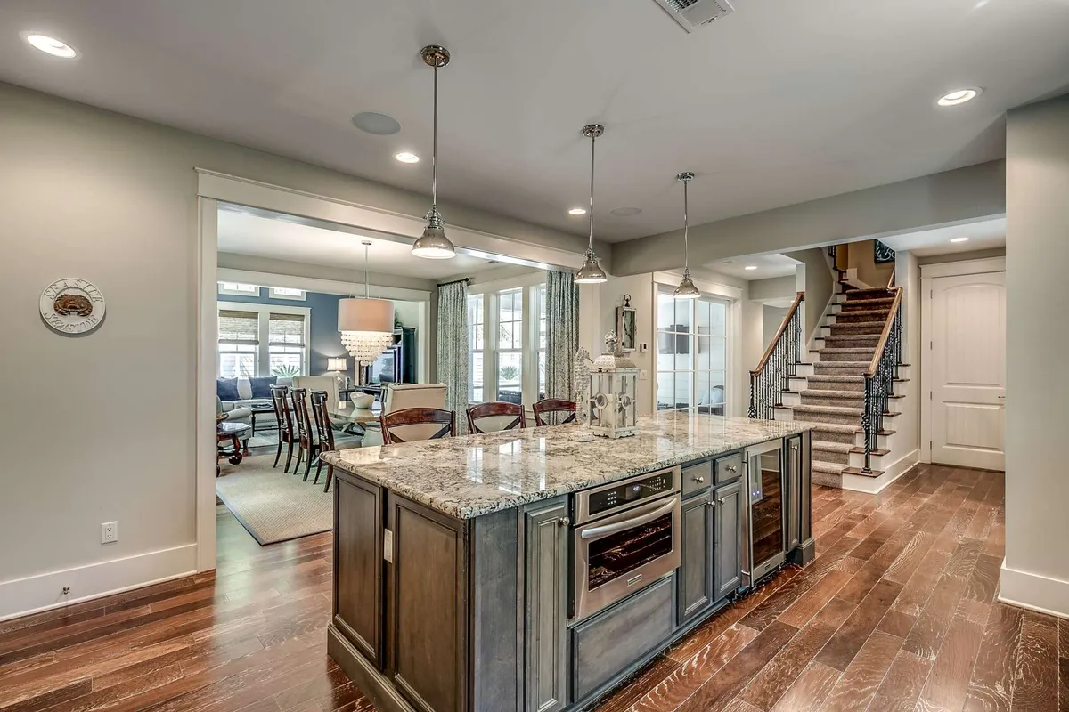 The kitchen island takes center stage, with elegant pendant lighting overhead and an open doorway leading to a connected living area. The granite countertops and modern fixtures create a sense of continuity between cooking, dining, and relaxing spaces.