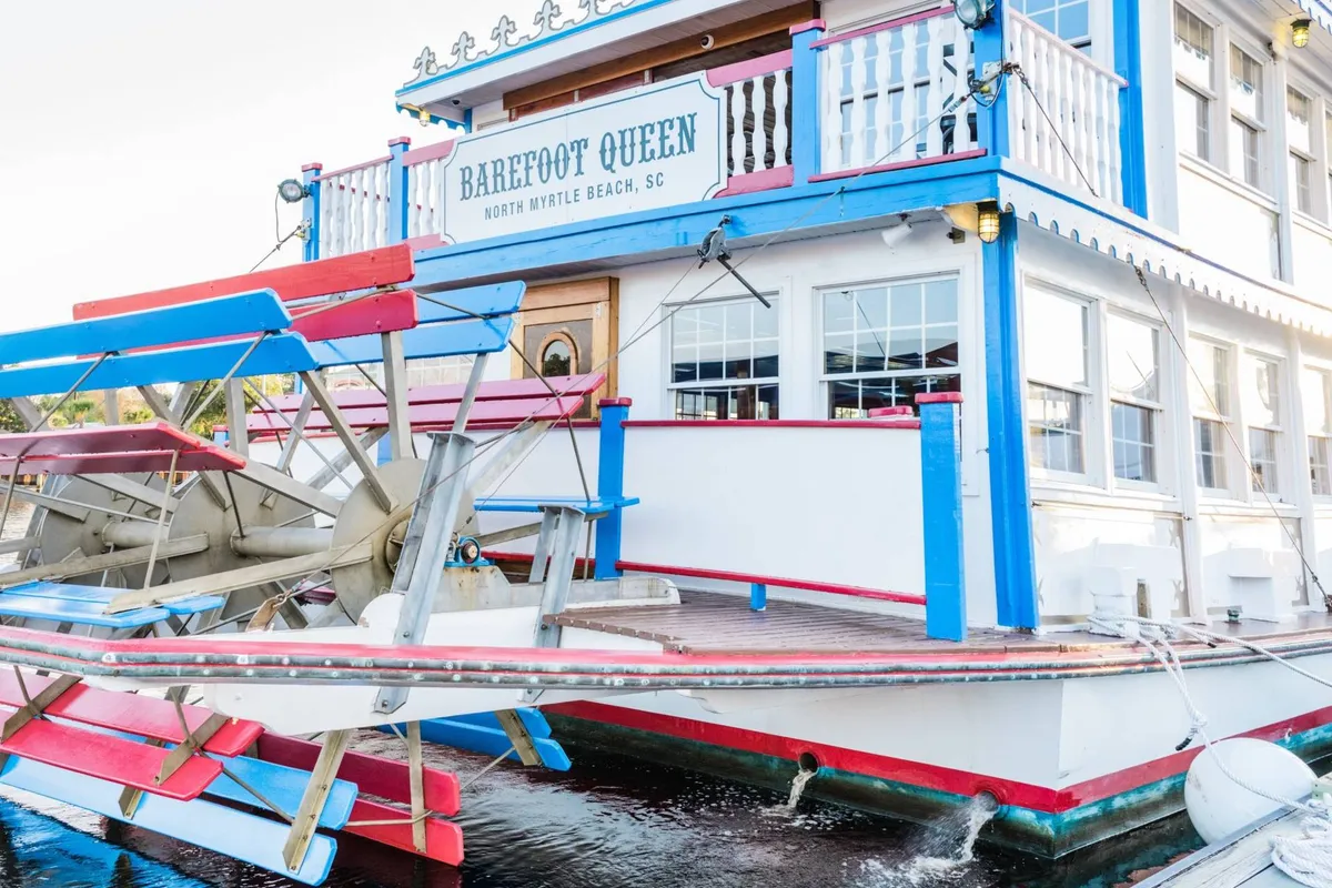 A cheerful exterior shot of the 'Barefoot Queen' paddlewheel riverboat with colorful signage, decorative railings, and the open sky. The scene suggests a fun outing, with opportunities for sightseeing, dining, and themed events on the water.