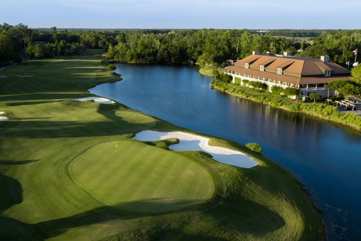 A sweeping exterior view of a lush golf green bordered by sand traps, a gleaming lake, and the elegant clubhouse in the background. The scene captures the quintessential golf experience, with manicured fairways and a tranquil setting ideal for both play and relaxation.
