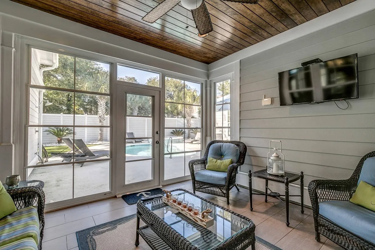 A bright, airy living area featuring spacious, comfortable seating arranged to foster conversation and relaxation. Large windows offer a view of the pool, and a wall-mounted television and decorative ceiling fan add both functionality and style. The environment is set up for social activities such as game nights, movie watching, or unwinding after a day out.