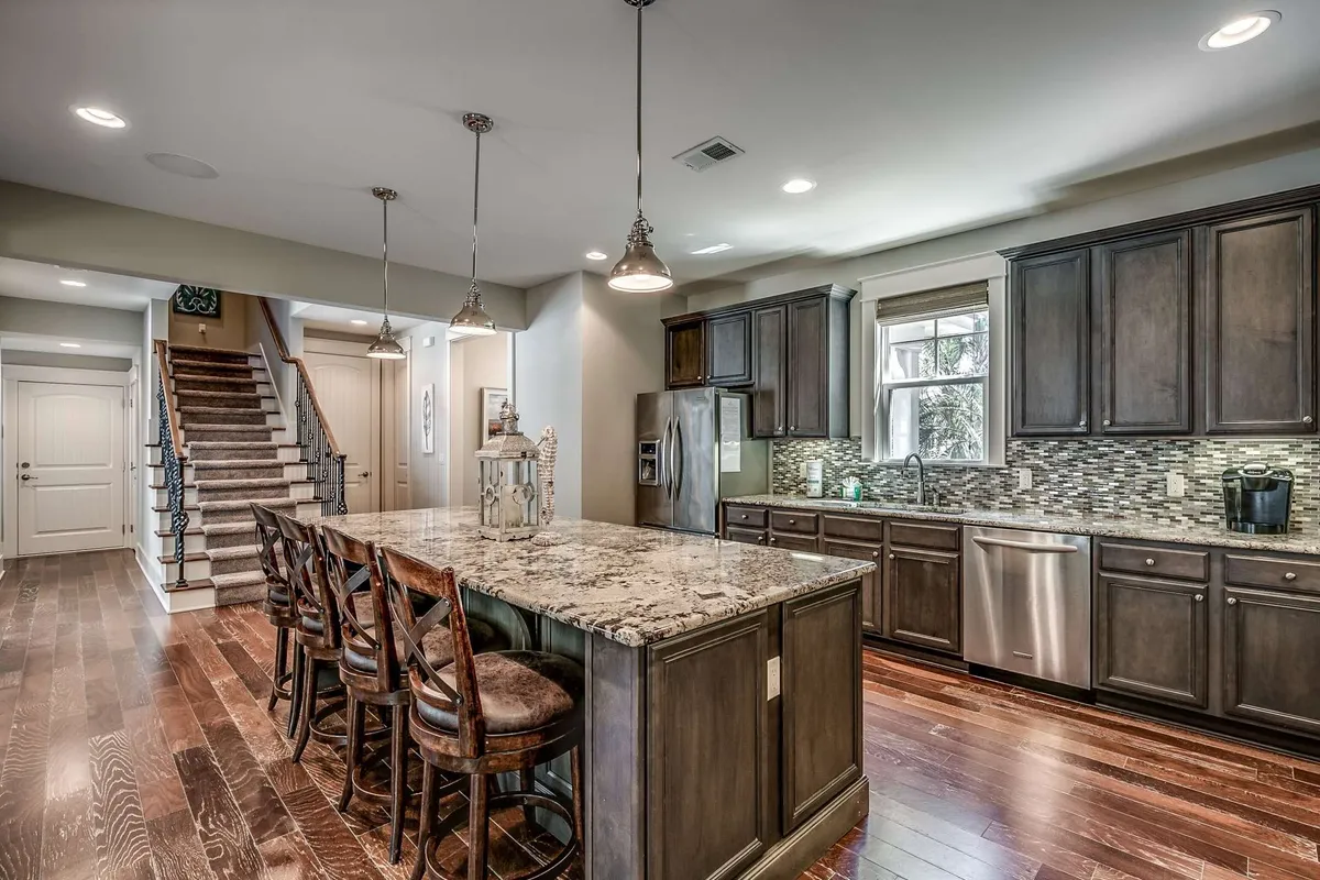 A view of the kitchen with a prominent granite island, wooden bar stools, and a suite of stainless steel appliances. The space is brightly illuminated by pendant lights, and the layout encourages social interaction whether cooking, dining, or enjoying coffee together.